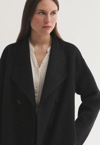 Femme aux longs cheveux bruns portant un manteau noir croisé par-dessus une chemise blanche boutonnée, regardant sur le côté sur un fond uni.