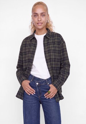 Une femme porte une chemise à carreaux foncés par-dessus un haut blanc et un jean bleu. Elle se tient devant un fond blanc uni.