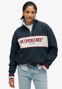 Sudadera navy con una franja blanca y roja que presenta "SUPERDRY". Cuello alto, cierre con cremallera, puños acanalados, colocada sobre una camisa de color azul claro.