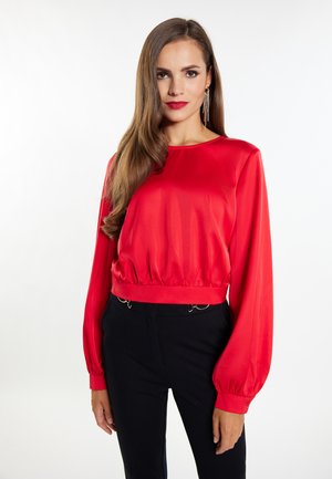 Femeie cu păr lung și șaten, purtând o bluză roșie cu mâneci lungi și pantaloni negri, stând în fața unui fundal alb simplu.