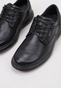 Par de zapatos casuales negros de cuero con cordones, con superficie texturizada y suelas de goma sobre un fondo claro.