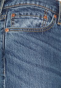 Vaqueros de mezclilla azul con una superficie texturizada, que presentan remaches de cobre, costuras naranjas y un pequeño bolsillo delantero. Tienen un ajuste ceñido.