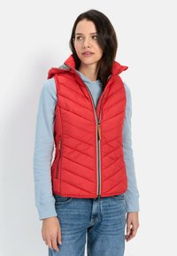 Rød quiltet vest med lynlås, hætte og sidelommer, lagdelt over en lyseblå skjorte med lange ærmer, parret med blå jeans.