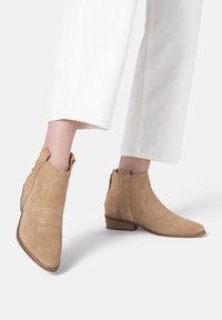 Botines de ante en beige con costuras decorativas y punta afilada. Cuentan con un tacón bajo apilado y cremalleras laterales para facilitar su uso.