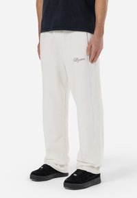 White sweatpants with subtle logo and side piping, featuring a relaxed fit and slightly textured fabric, paired with black sneakers.