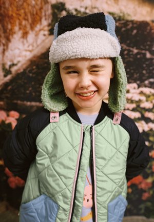 Child wearing a colorful winter hat and quilted jacket smiles and winks with hands behind their back in front of a floral background.