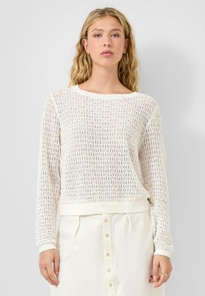 Blondynka z kolczykami-kołami, ubrana w biały, dziany sweter z długim rękawem oraz białą, zapinaną na guziki spódnicę z wysokim stanem.