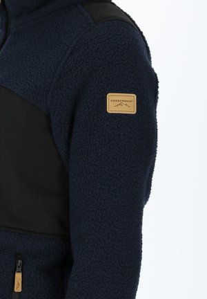 Giacca in pile blu navy con dettagli in poliestere nero. Presenta una finitura testurizzata, tasca laterale con zip e un'etichetta in tessuto color sabbia sul manica.