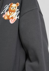 Sudadera gris con un gráfico de oso impreso en marrón y rosa, con una textura suave y un diseño de corte relajado.
