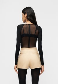 Γυναί κα με μακριά σκούρα μαλλιά, που φορά διαφανές μακρυμάνικο μαύρο μπλουζάκι, σορτς μπεζ με ψηλή μέση και μαύρες καλσόν, γυρισμένη πλάτη σε λευκό φόντο.