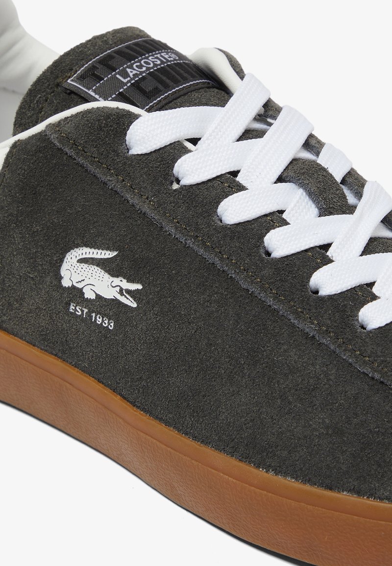 Sneakers Lacoste z szarym zamszowym wierzchem, białymi sznurówkami i brązową gumową podeszwą. Posiadają logo krokodyla oraz szczegół "EST 1933".