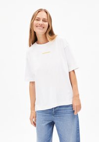 Lächelnde Frau, die ein weißes, übergroßes T-Shirt mit dem Text "ARMEDANGELS" und hellblauen Jeans trägt, vor einem schlichten weißen Hintergrund steht.