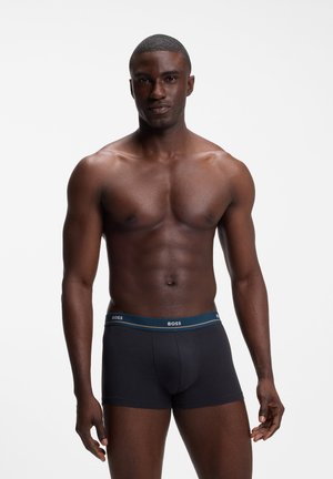 Muskelmand med mørk hud står med front mod kameraet iført sorte BOSS boxer shorts foran en ensfarvet hvid baggrund.
