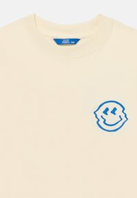 Kremowy T-shirt z niebieskim uśmiechniętym logo na lewej piersi oraz metką "REBEL 140" wewnątrz kołnierza.