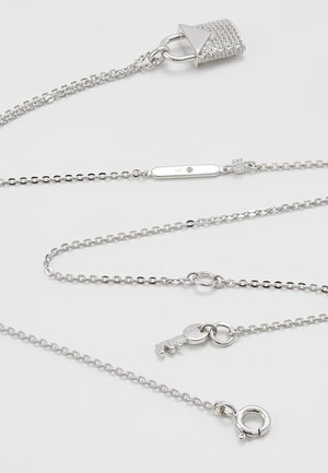 Chaînes en argent avec un petit pendentif clé, un pendentif cadenas incrusté de cristaux et un fermoir sur un fond clair.