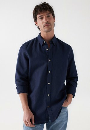 Camisa azul marino de lino con botones, mangas largas y corte holgado. Cuenta con cuello y botones color beige en la parte frontal.