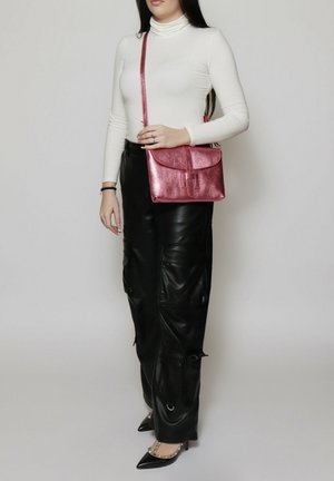 Sac à épaule métallique rose avec un rabat et une sangle ajustable, porté avec un col roulé blanc ajusté et un pantalon cargo en cuir noir.