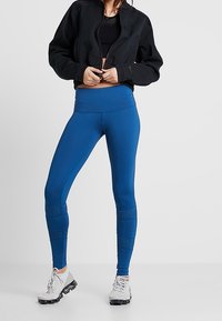 Blå leggings med hög midja, med strukturerade detaljer nära fållen och en slät yta. Bärs med en svart kort jacka och ljusa sneakers.