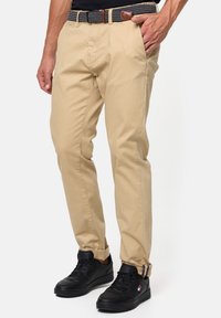 Pantalones chinos beige de corte slim, con bolsillos laterales y puños enrollados, combinados con zapatillas negras y un cinturón de estampado.