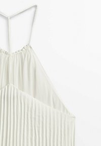 Haut blanc sans manches avec bretelles fines, encolure froncée et plis verticaux sur la partie inférieure, suspendu à un cintre blanc.