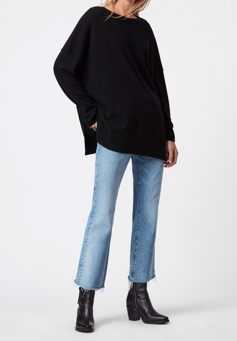 Personne portant un pull noir oversize, un jean court bleu clair avec des bords effilochés, et des bottines noires à talons, se tenant devant un fond blanc.