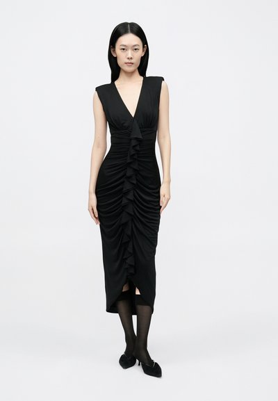 Fekete, testhez simuló ruha mély V-nyakkivágással; függőleges ráncokkal, a középen fodrokkal és térdig érő hasítékkal. A modell áttetsző fekete harisnyát és cipőt visel.