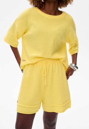 Kobieta ubrana w luźny, teksturowany, żółty sweter z krótkim rękawem oraz dopasowane szorty z troczkiem, z srebrnymi bransoletkami na prawym nadgarstku.