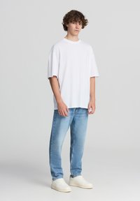 T-shirt bianca in cotone con una vestibilità rilassata, maniche corte e collo rotondo, abbinata a jeans dritti blu chiaro e sneakers bianche.