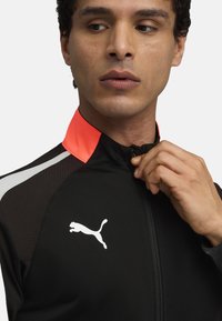 Giacca sportiva nera con colletto alto, presenta un accento rosa sul colletto, strisce bianche e un logo bianco sul petto. Chiusura con zip frontale.