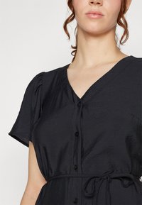 Schwarzes Kleid mit V-Ausschnitt, kurzen Puffärmeln, Knöpfen vorn und einem Bindegürtel. Der Stoff hat einen leichten Glanz und eine plissierte Textur.