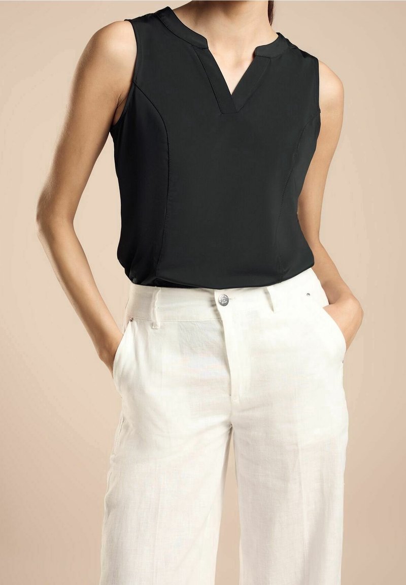 Femme portant un haut noir sans manches à col en V et un pantalon blanc taille haute, les mains dans les poches, sur fond beige.