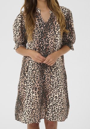 Kvinde iført en løs, knælængde kjole med leopardprint og trekvart puffede ærmer, hænderne foldet foran.