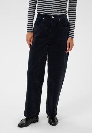 Personne portant un pantalon large noir, des mocassins noirs et un haut rayé gris et noir à manches longues sur fond uni.