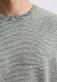 Gros plan sur une personne portant un pull en maille texturée vert clair avec un col rond côtelé et un logo brodé subtil sur la poitrine.