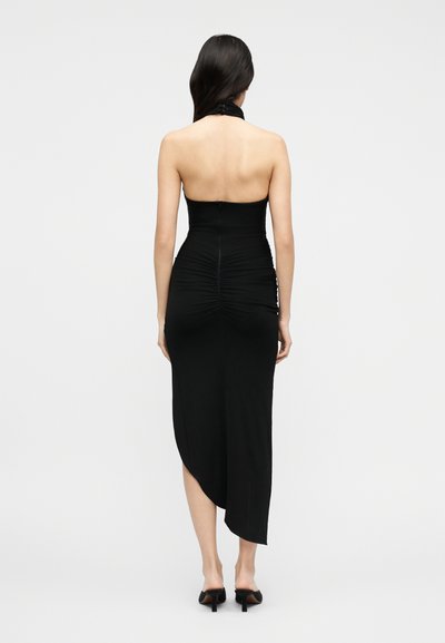 Γυναίκα με μακριά σκούρα μαλλιά που φοράει μαύρο ασύμμετρο μίντι φόρεμα χωρίς τιράντες και μαύρα mule με τακούνι, που στέκεται μπροστά σε λευκό φόντο.