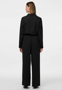 Črn kombinezon z dolgimi rokavi in manšetami. Ima elastičen pas in široke hlače ter je izdelan iz teksturirane tkanine.