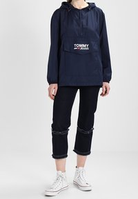 Námorně modrá lehká anoraková bunda s kapucí a přední kapsou, s bílým logem "TOMMY JEANS", v kombinaci s tmavými zkrácenými džíny a bílými teniskami.