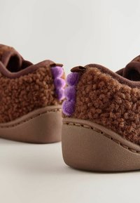 Chaussons marron doux avec des détails en pom-pom violet à l'arrière et une semelle texturée robuste sur fond blanc.