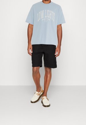 Lyseblå bomuldst-shirt med "LOW LIGHTS" grafisk tryk, kombineret med sorte shorts og beige sandaler med stropdesign.