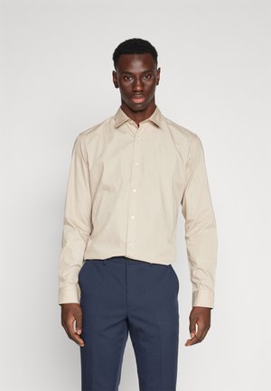 Chemise beige à manches longues avec un col pointu, devant à boutons et boutons blancs. Le tissu semble lisse avec un léger éclat, portée avec un pantalon bleu.
