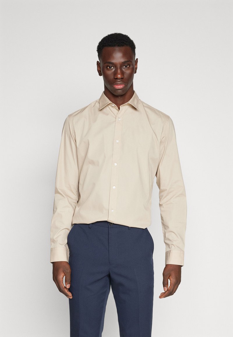 Beige Langarmhemd mit spitzem Kragen, Knopfverschluss und weißen Knöpfen. Der Stoff wirkt glatt mit leichtem Glanz und wird mit blauen Hosen getragen.