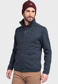 Navyblaue Zip-Jacke mit strukturiertem Muster, hohem Kragen und kontrastierendem Reißverschluss. Kombiniert mit einer burgunderroten Strickmütze.