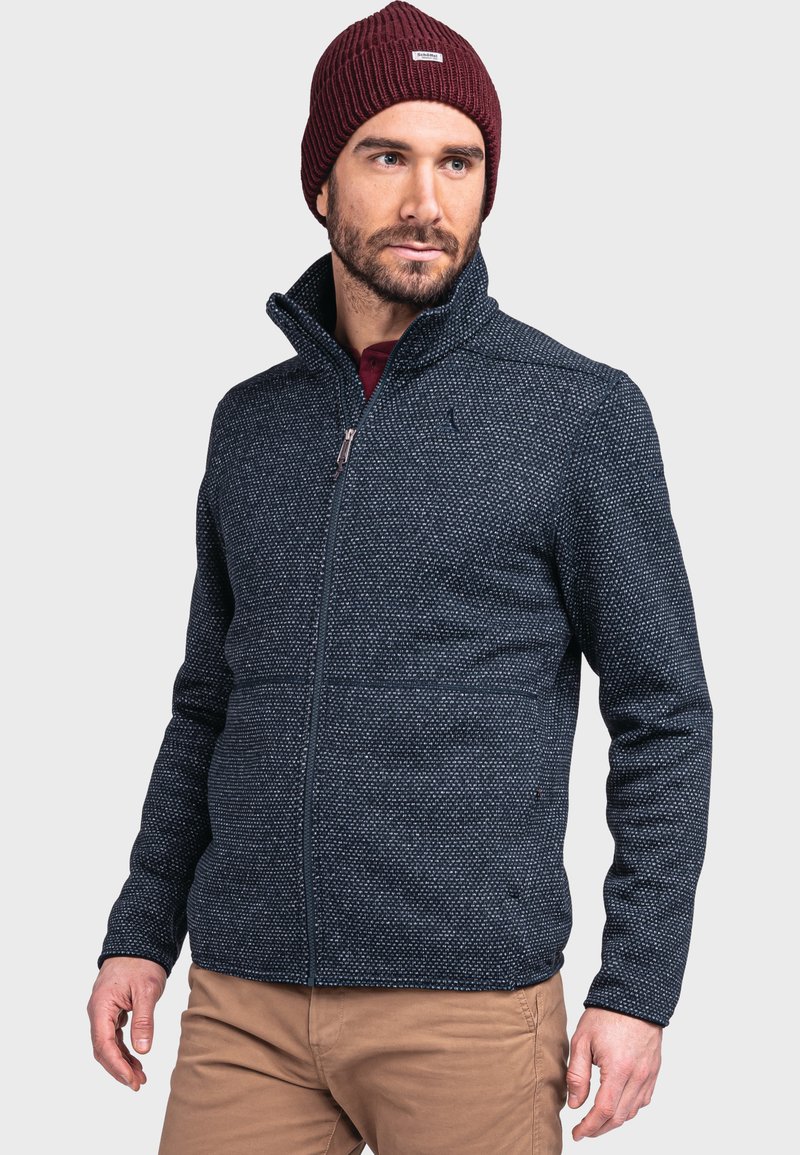 Navyblaue Zip-Jacke mit strukturiertem Muster, hohem Kragen und kontrastierendem Reißverschluss. Kombiniert mit einer burgunderroten Strickmütze.