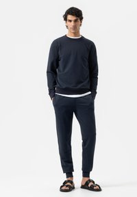 Man staat met zijn handen in zijn zakken, gekleed in een marineblauwe sweatshirt en joggingbroek, met zwarte sandalen, tegen een witte achtergrond.