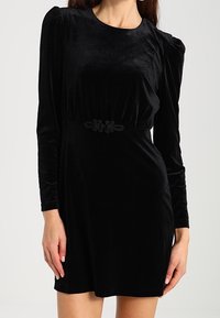 Vestido de terciopelo negro con mangas largas, escote redondo y hombros abullonados. Presenta un cinturón decorativo negro en la cintura. Textura suave.