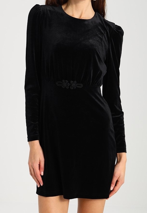 Vestido de terciopelo negro con mangas largas, escote redondo y hombros abullonados. Presenta un cinturón decorativo negro en la cintura. Textura suave.