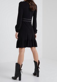 Robe noire à manches longues avec un ourlet froncé et une ceinture cloutée. Associée à des bottes noires jusqu'au genou avec un détail à nouer à l'arrière.