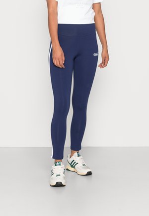 Leggings navy de material liso y elástico con rayas blancas en los laterales y una cintura alta. Combinados con zapatillas deportivas blancas y verdes.