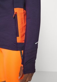 Top de manga larga púrpura con bolsillo lateral naranja y detalles reflectantes. Tela texturizada, diseño ceñido. Pantalones cortos naranjas visibles en la parte inferior.