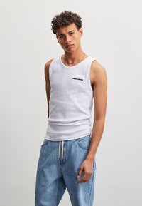 Jeune homme aux cheveux bouclés porte un débardeur blanc avec le texte "PEGADOR" et un jean bleu ample, debout devant un fond clair et uni.